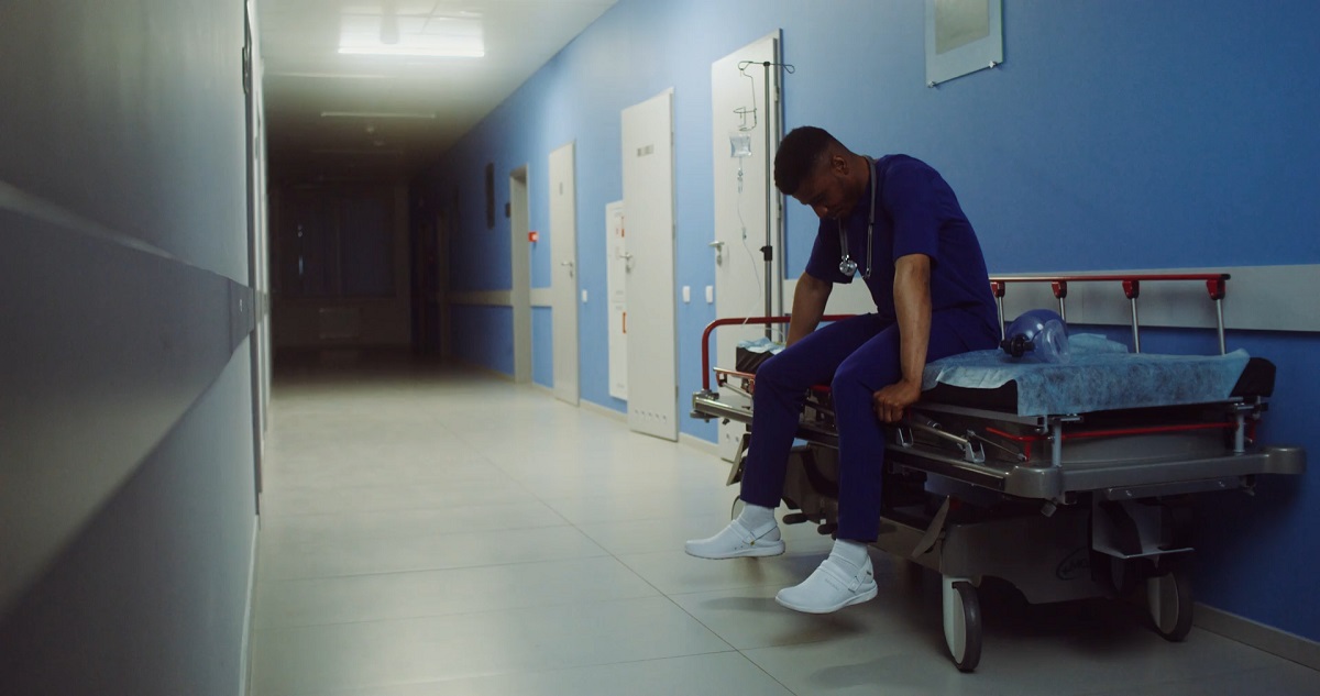 A tired doctor in blue scrubs sitting on a hospital stretcher in a dimly lit hallway